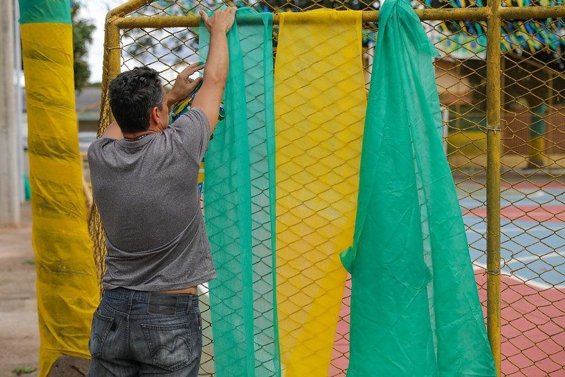 Homem branco de camisa cinza de costas pendurado bandeiras verde e amarelo em quadra poliesportiva