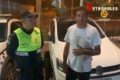 Policial militar com farda verde fluorescente e azul-escuro, usando boné azul-escuro da corporação, ao lado do homem pardo de camiseta branca, vem em frente a carro da PMDF e um Volkswagen branco