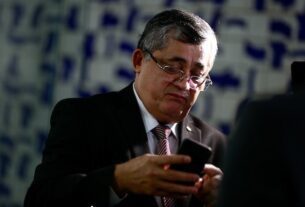 O deputado petista e membro da equipe de transição do governo Lula, José Guimarães. Ele usa óculos, olhando e mexendo em celular com mural da Câmara ao fundo - Metrópoles